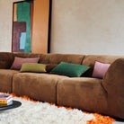 Een bruine pluche sofa met HK Living Kussen Country House Groen 60/35 kussens rust op een wit shagtapijt. Een abstract schilderij leunt tegen een beige muur en op een zwarte salontafel staan boeken.