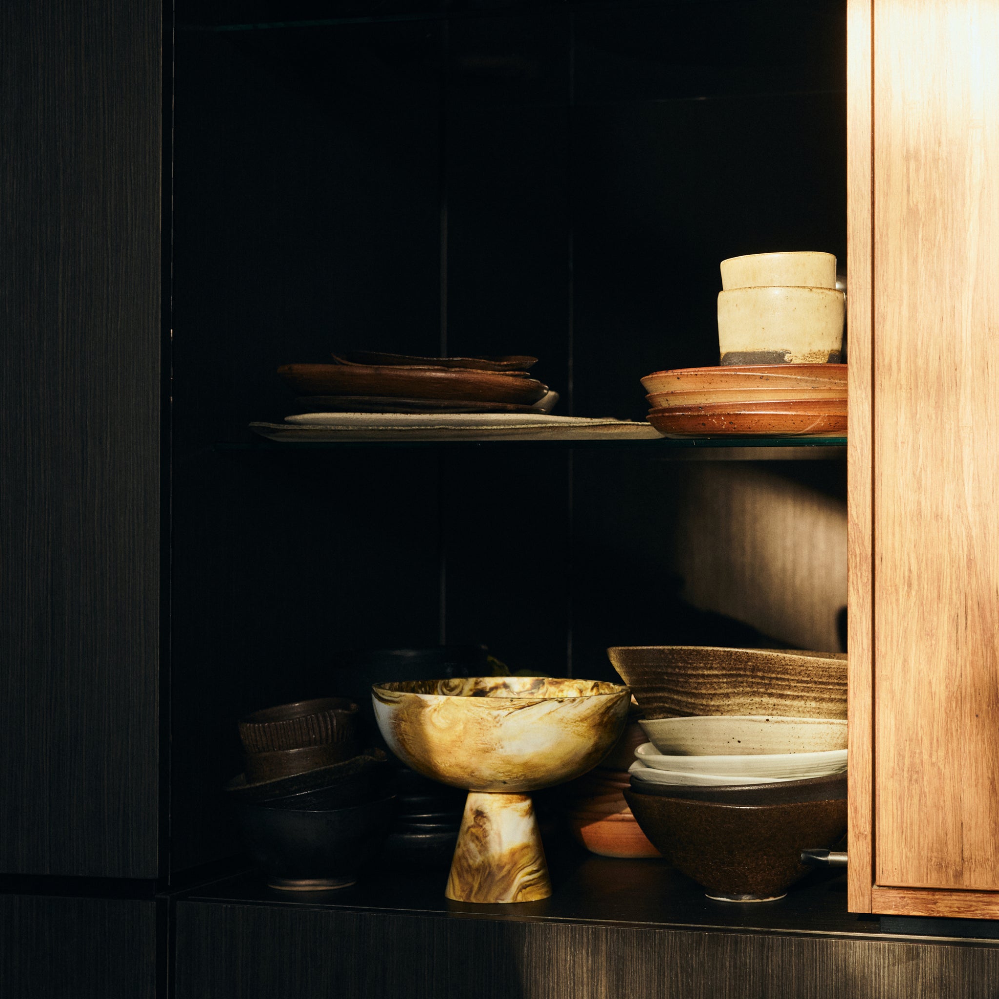 Een schemerig verlichte houten kast met open planken toont een assortiment keramische HK Living kommen, borden en kopjes in aardetinten, waaronder de opvallende HK Living gemarmerde Kom op Voet Oker D23, verlicht door een warme zonnestraal.