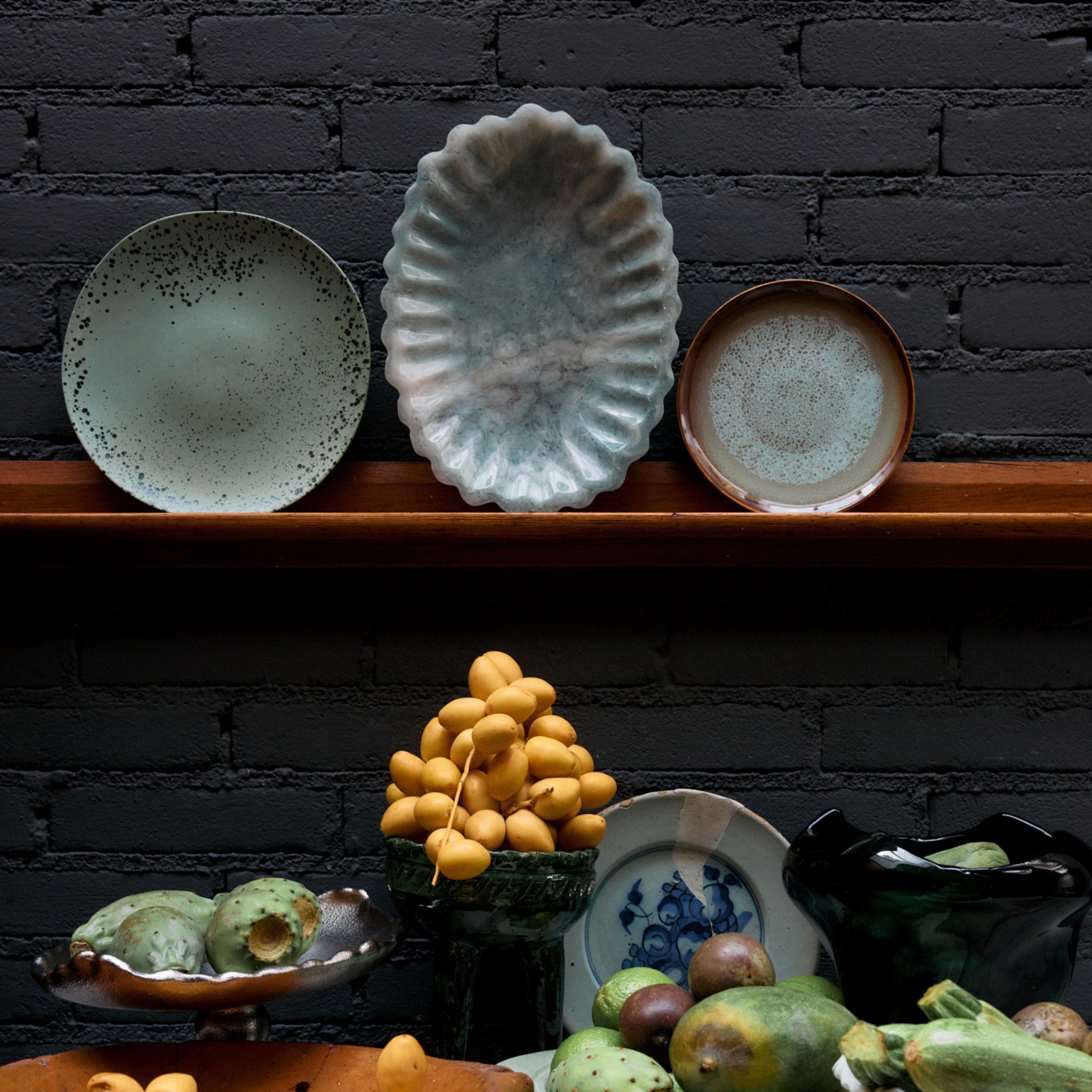 Drie decoratieve borden op een houten plank tegen een donkere bakstenen muur, met de HK Living New Classics Resin Schaal Lichtblauw en diverse groene vruchten op de tafel eronder, die moderne elegantie toevoegen.