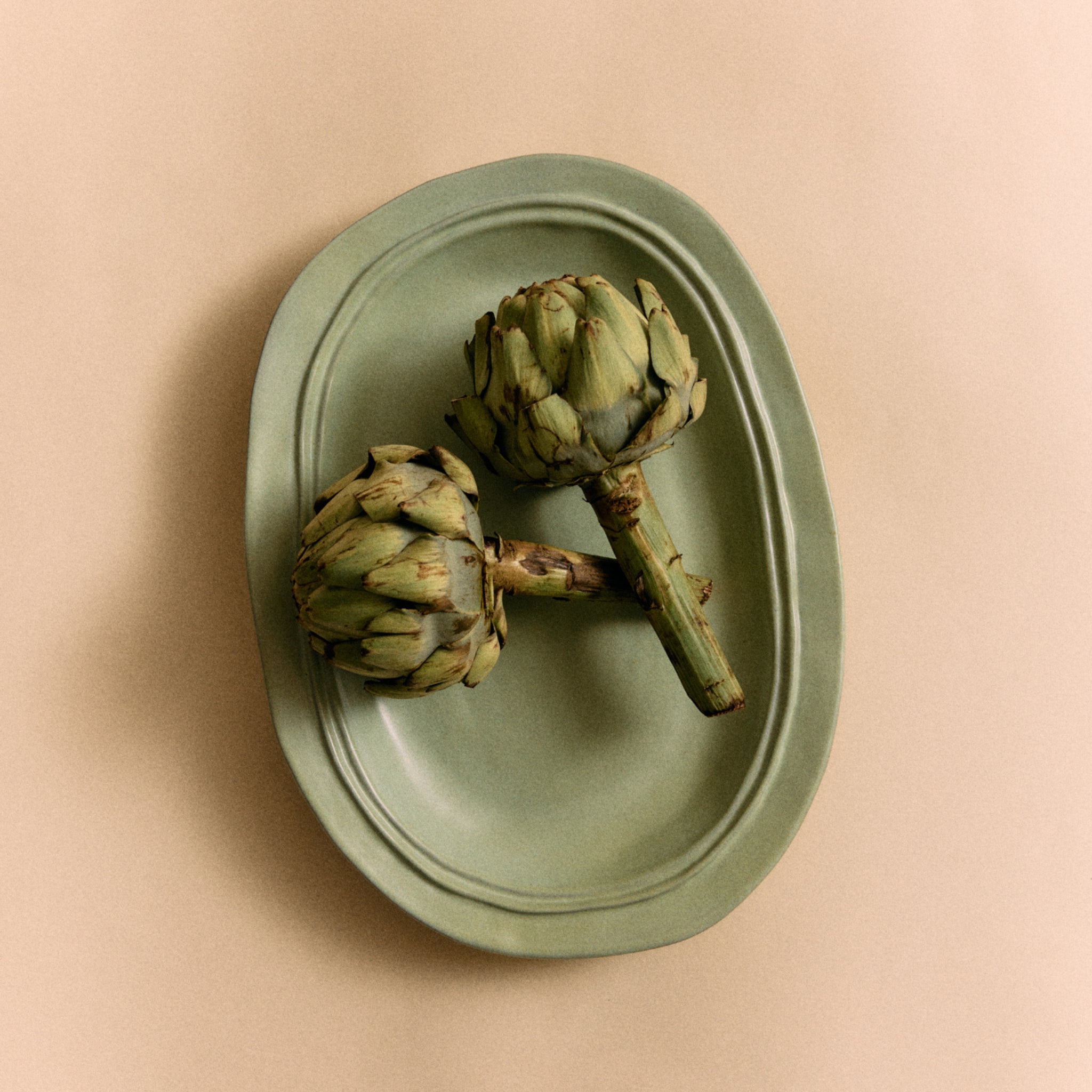 Twee verse artisjokken rusten op een HK Living New Classics Schaal Muntgroen L, onderdeel van de keramische serviescollectie van HK Living, tegen een beige achtergrond.
