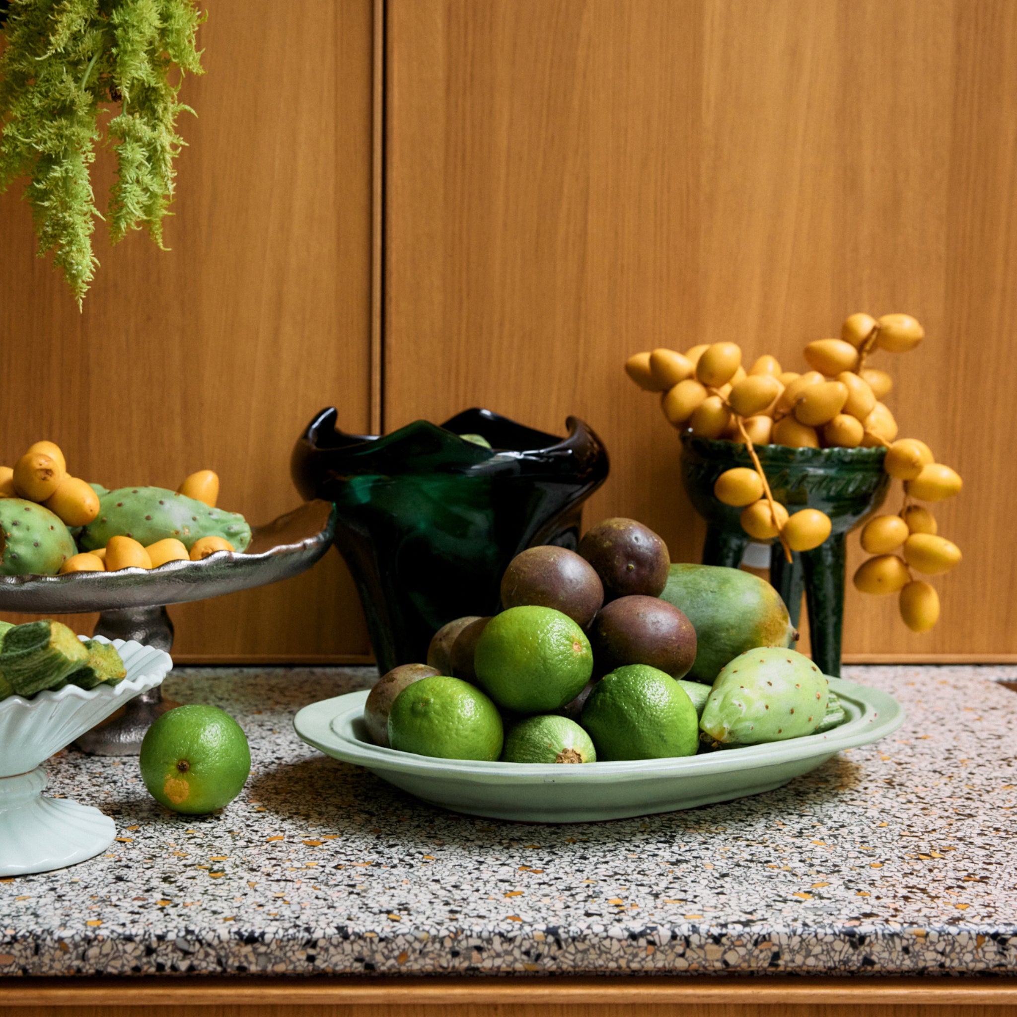 Een aanrecht toont groen en geel fruit - limoenen, cactusvijgen, dadels en passievruchten - gerangschikt in de HK Living New Classics Schaal Muntgroen L. Daarboven hangt groen met een donkere vaas tegen houten kasten.