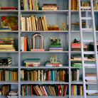 Een boekenplank met boeken in alle maten en kleuren, aangevuld met decoratief HK Living Paper Object: Naar Constructivisme en kunstwerken. Een rollende ladder leunt vlakbij en draagt bij aan de stijlvolle, eclectische sfeer van dit moderne interieur.