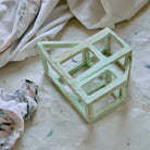 Het HK Living Paper Object: After Constructivism - een lichtgroene geometrische papier-maché sculptuur - rust op een witte, met verf besmeurde stof naast een verfrommeld doek, met gedrapeerde witte stof als achtergrond, perfect voor moderne interieurs.