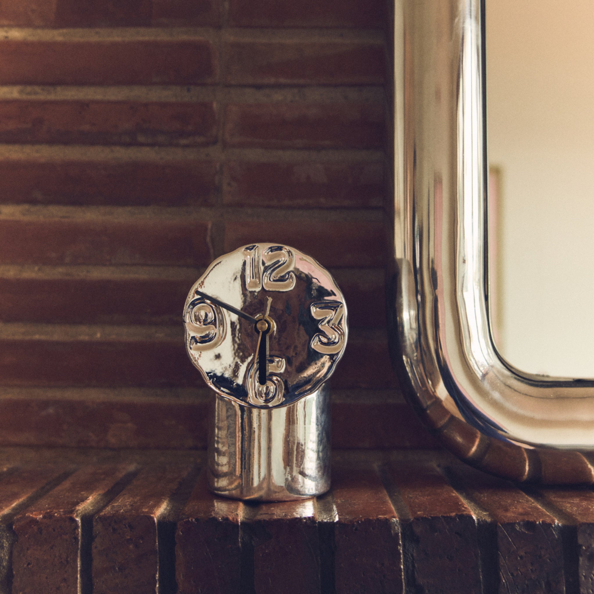 Een HK Living Retro Klok Chroom met een gesmolten, zilverkleurig ontwerp staat op een bakstenen schouw naast een spiegel met metalen frame en een organische vaas en creëert een warme, vintage sfeer.
