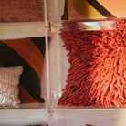 Een close-up van sierkussens op planken, met de HK Living Rugged Kussen Kersenrood 50/50, een glanzend zilver kussen, en HK Living kussens met warme geometrische patronen.