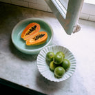 Een gehalveerde papaja ligt in de HK Living Saladekom Mint D26, terwijl vijf limoenen in een witte kom op een marmeren aanrecht staan bij een open raam waar het zonlicht naar binnen schijnt.