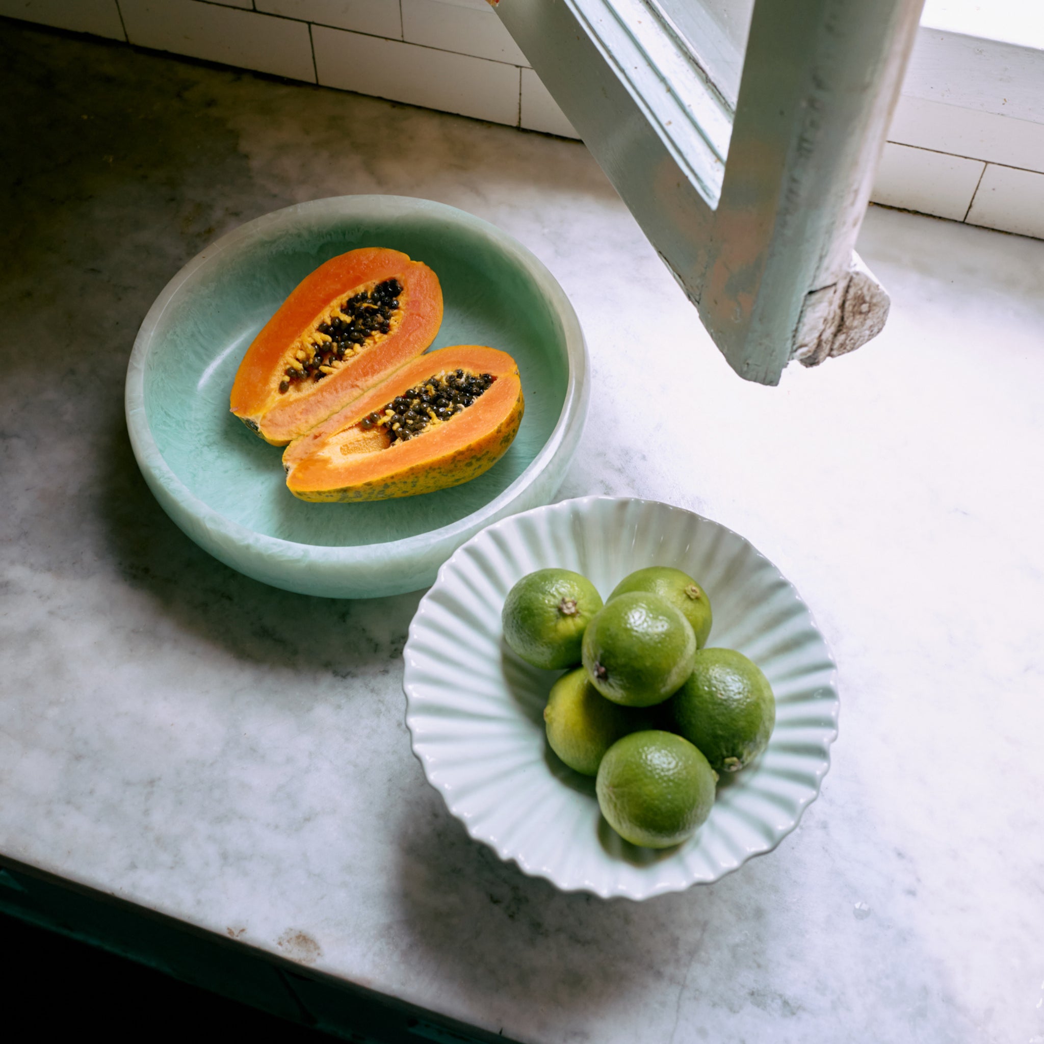 Een gehalveerde papaja ligt in de HK Living Saladekom Mint D26, terwijl vijf limoenen in een witte kom op een marmeren aanrecht staan bij een open raam waar het zonlicht naar binnen schijnt.