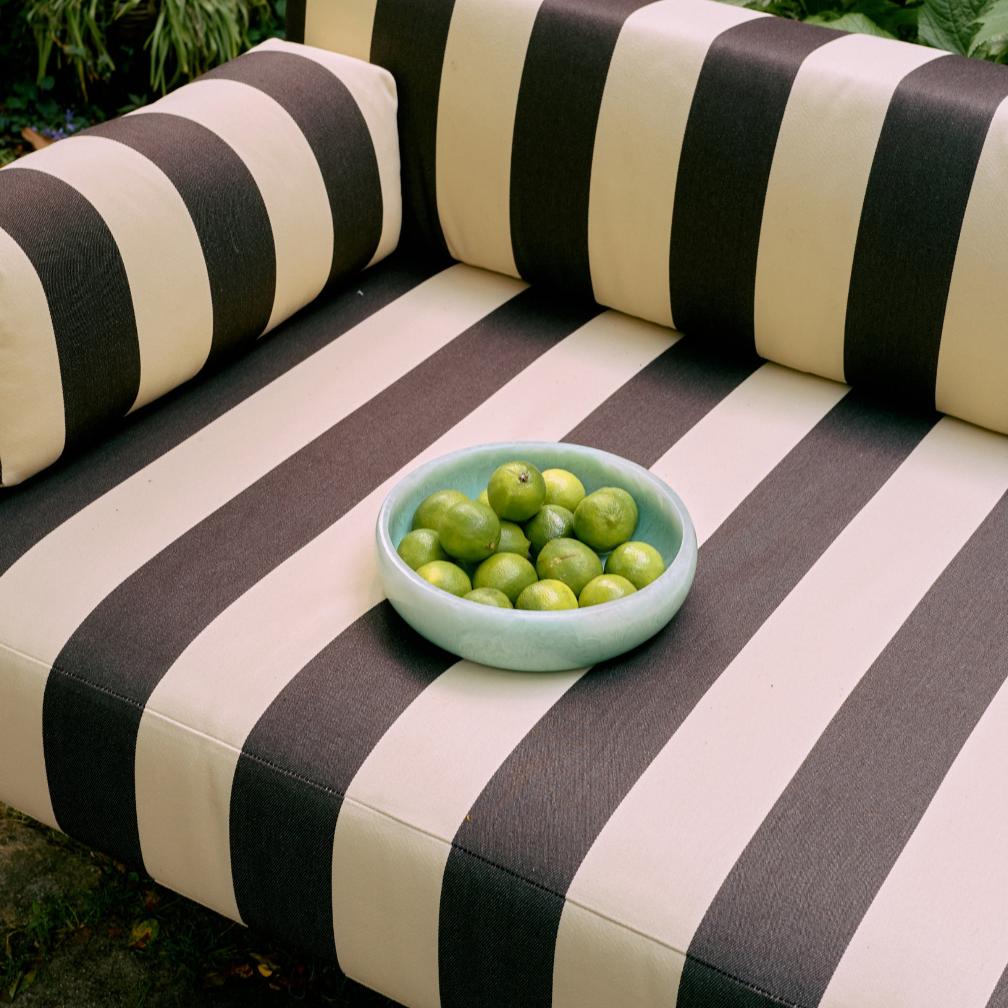 Een schaal met groene limoenen staat in een HK Living Saladekom Mint D26 van HK Living op een beige en donkerbruin gestreepte buitenbank, omringd door groen.