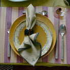 Een stijlvolle tafel is voorzien van een geel bord, een gedessineerd bijzetbord, een gevouwen servet met de HK Living Servetring Sunrise 4 st., zilverwerk op een gestreepte placemat en een helder glas voor een eigentijdse tafeldekking.