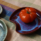 Een grote, rijpe tomaat ligt op de HK Living Shell Schaal Sienna, een kom van organisch keramiek van HK Living, geplaatst op een veelkleurige geruite doek naast een groen geglazuurd bord.