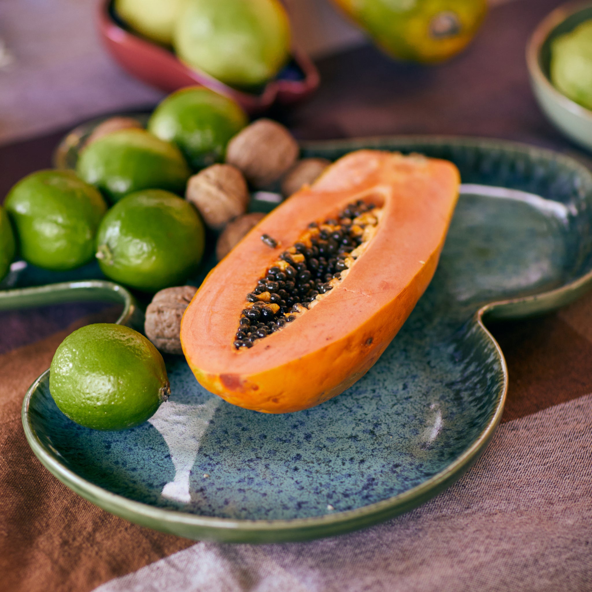 Een gehalveerde papaja met zwarte zaden staat op een HK Living Schaal Schaal Teal, omringd door hele limoenen en walnoten op een tafelkleed met patroon.