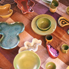 Kleurrijke keramische borden, kommen en kopjes, waaronder een blauwgroen servies en de HK Living Shell Schaal Teal, zijn gerangschikt op een geruit bruin en paars tafelkleed, waardoor een levendige en artistieke tafelsetting ontstaat.