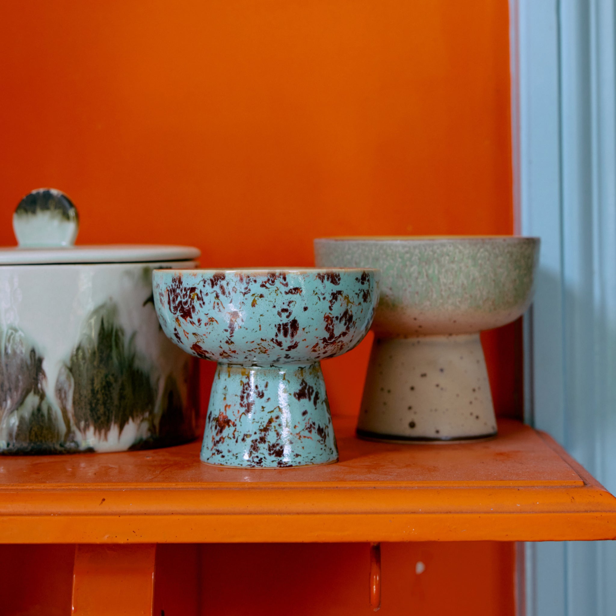 Drie HK Living 70s Ceramics Tapas Kom op Voet Patina S kommen hebben unieke gespikkelde en gemarmerde ontwerpen in blauwe, groene en neutrale tinten. Ze staan op een oranje plank met een oranje en lichtblauwe muur als achtergrond.