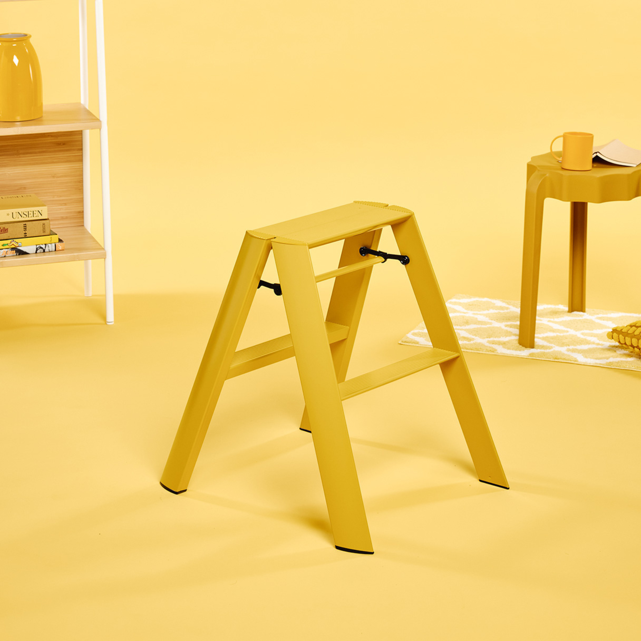 Een Hasegawa Lucano Ladder 2 Step Geel van Hasegawa staat in een kamer waar de muren, de vloer en het meubilair – waaronder een tafeltje met een kopje en boeken op een plank – allemaal geel zijn, waardoor een monochroom geel thema ontstaat dat is geïnspireerd op Japans design.