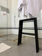 De Hasegawa Lucano Ladder 2 Step Zwart van Hasegawa, een strakke, zwarte, compacte huishoudtrap, staat op een betonnen badkamervloer waarop witte handdoeken zijn neergelegd. De trap staat naast een glazen douchecabine met een wit betegelde wand, een douchekop en een wit badmatje, en vertoont subtiele invloeden uit het Japanse design.