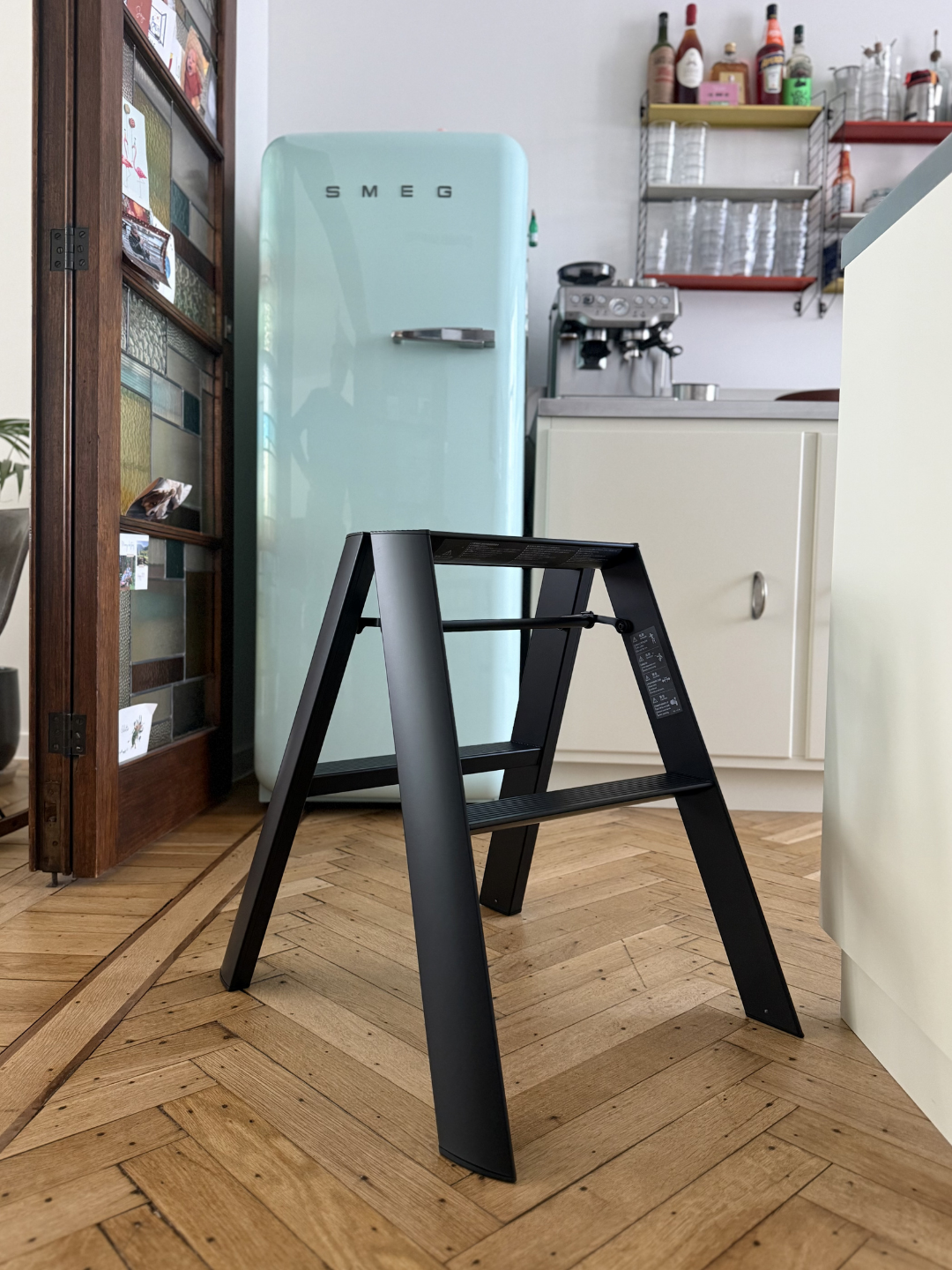 Een compacte Hasegawa Lucano-ladder met twee treden in het zwart, uitgevoerd in een strakke zwarte afwerking, staat op een houten vloer in een keuken. Op de achtergrond zijn een lichtblauwe SMEG-koelkast, keukenkastjes en een koffiezetapparaat te zien.