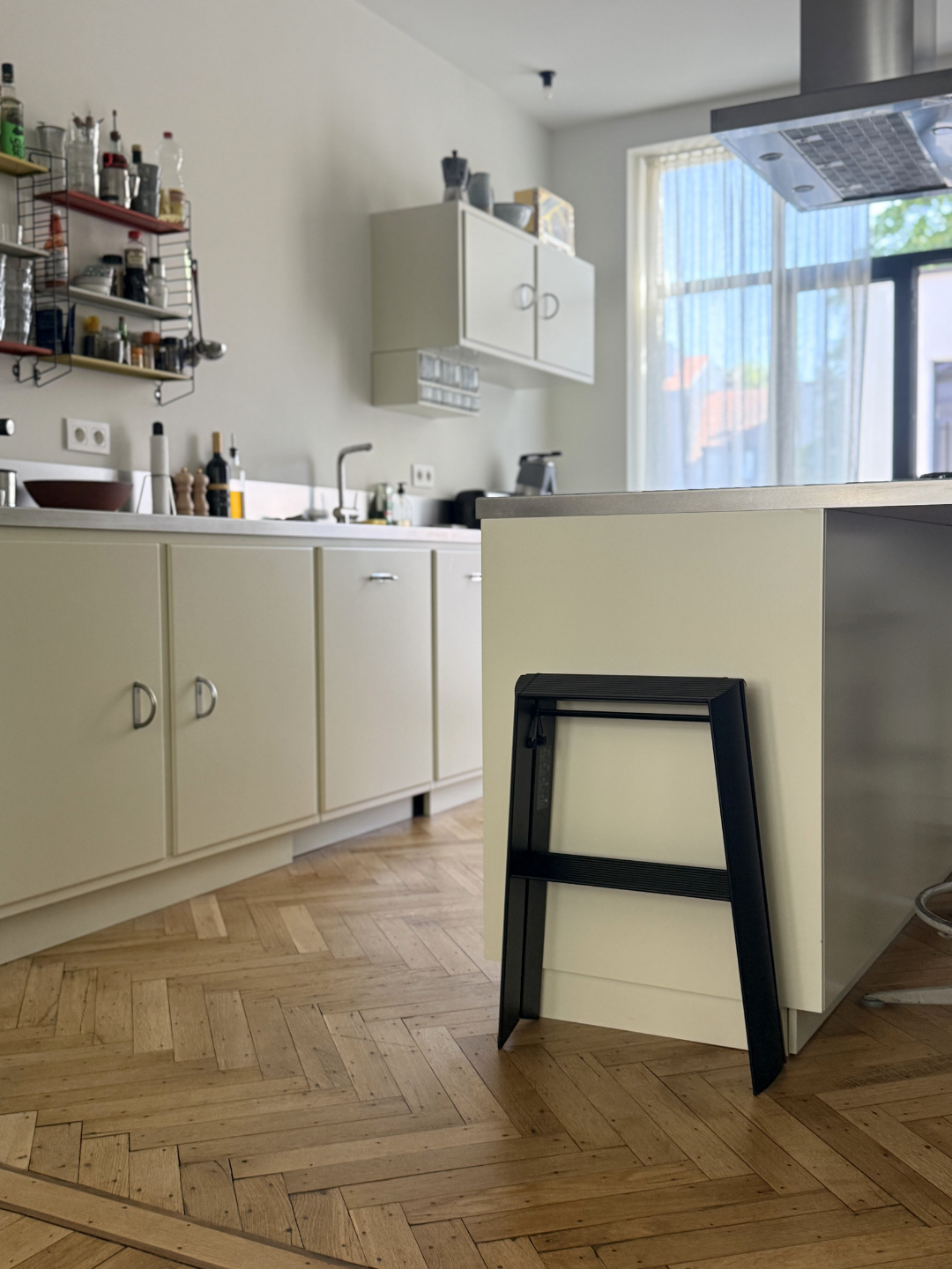 Moderne keuken met lichte kasten, een houten visgraatvloer, een strakke Hasegawa Lucano Ladder 2 Step Zwart van Hasegawa die tegen het kookeiland leunt, aan de muur bevestigde planken geïnspireerd op Japans design, en een groot raam dat natuurlijk licht binnenlaat.
