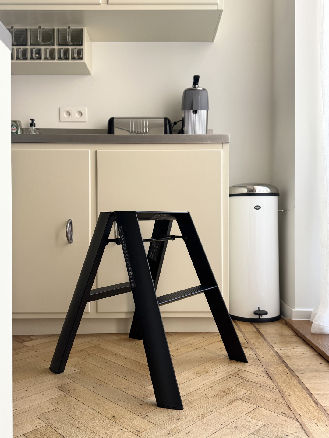 Een Hasegawa Lucano-ladder met twee treden in de kleur zwart, van Hasegawa, met een op Japan geïnspireerd ontwerp, staat op een houten keukenvloer voor crèmekleurige kasten en een vuilnisbak, met een koffiezetapparaat en een broodrooster op het aanrecht.