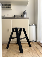 Een Hasegawa Lucano-ladder met twee treden in de kleur zwart, van Hasegawa, met een op Japan geïnspireerd ontwerp, staat op een houten keukenvloer voor crèmekleurige kasten en een vuilnisbak, met een koffiezetapparaat en een broodrooster op het aanrecht.
