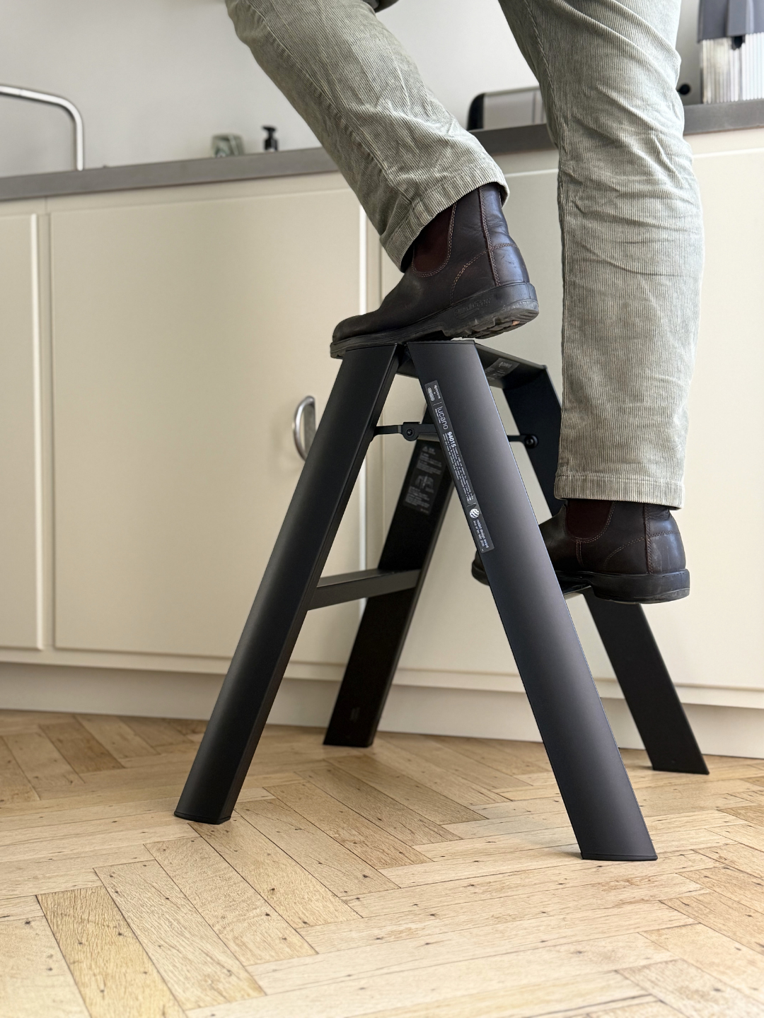 Een persoon met een lichtgroene broek en donkere laarzen staat op een Hasegawa Lucano Ladder 2 Step Zwart van Hasegawa in een keuken met een lichte houten vloer en crèmekleurige kasten.
