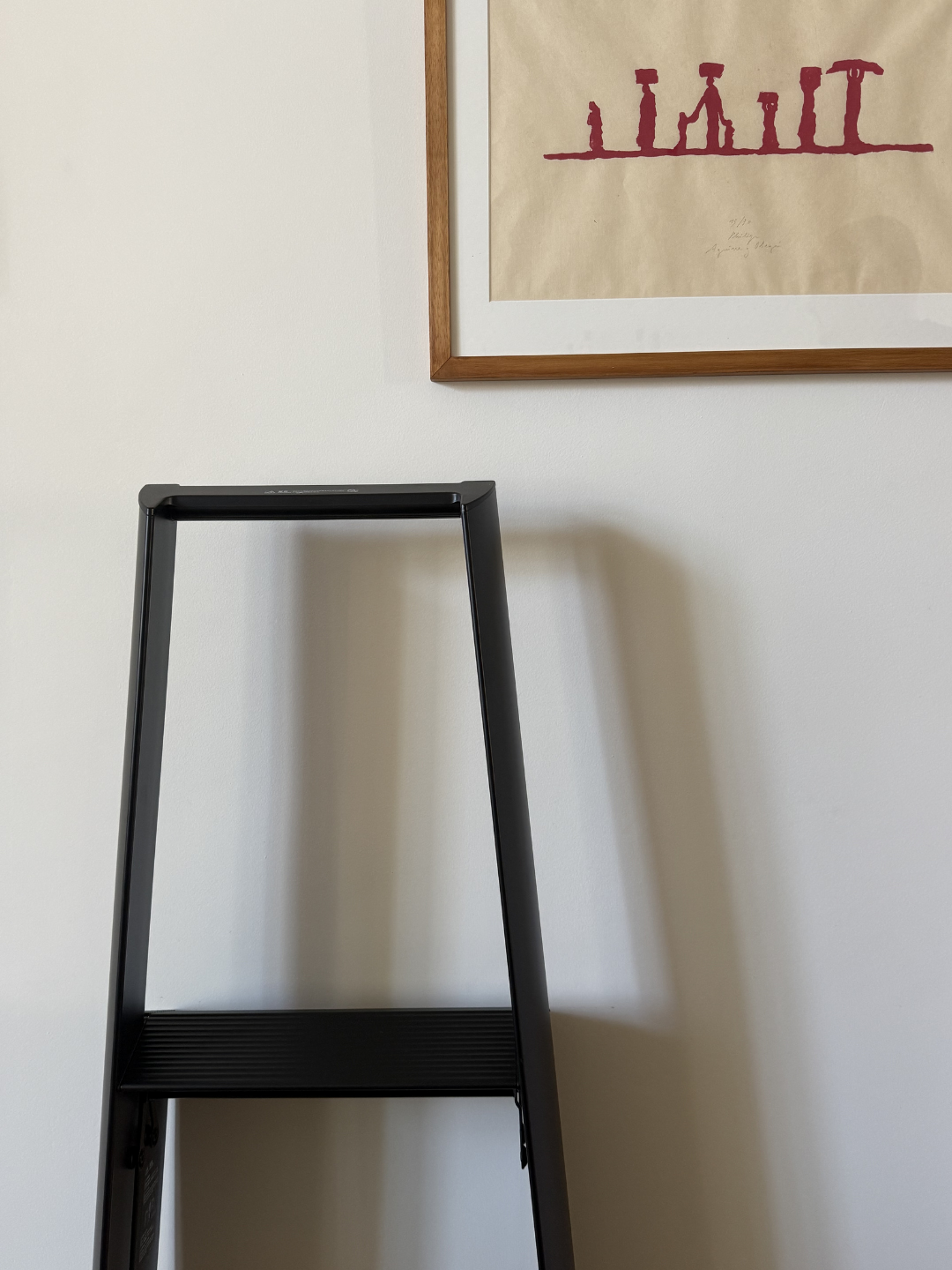 Een Hasegawa Lucano Ladder 3 Step Zwart van Hasegawa staat tegen een witte muur, onder een ingelijst kunstwerk versierd met rode abstracte vormen, waarmee de ingetogen invloeden van het Japanse design goed tot uiting komen.