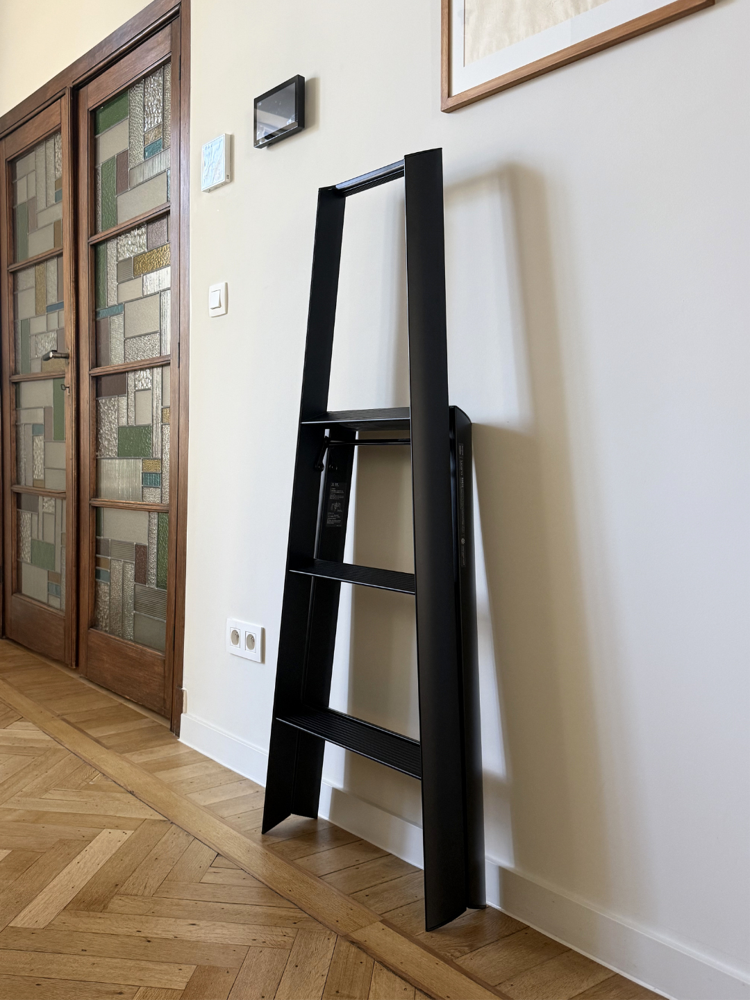 Een Hasegawa Lucano-ladder met drie treden in het zwart, van Hasegawa, met een strak, op Japanse stijl geïnspireerd ontwerp van zwart metaal, staat stijlvol tegen een lichtgekleurde muur in een gang met houten deuren en parketvloer.