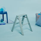 Een Hasegawa Lucano Ladder 2 Step in stoffig mint van Hasegawa staat op een lichtblauwe vloer, vergezeld door een blauwe bijzettafel met boeken en decor, een grijze zitkussens en een lichtblauw kussen in een minimalistische Japans geïnspireerde omgeving.