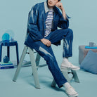 Een persoon in een blauw spijkerjack, spijkerbroek, gestreept shirt en witte sneakers zit op een Hasegawa Lucano Ladder 1 Stap Dusty Mint in een moderne, blauw gethematiseerde kamer met minimale inrichting, boeken en strakke designaccenten.