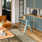 Moderne leefruimte met houten en blauwe kasten, een bruine leren bank, lichthouten salontafel en beige vloerkleed. De Hasegawa Lucano Ladder 2 Step in Dusty Mint staat in het middelpunt van de belangstelling en benadrukt het minimale Japanse ontwerp. Deuropening onthult bureau en stoel.