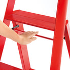 Een hand raakt de horizontale steunbalk aan van de Hasegawa Lucano Ladder 3 Step Rood van Hasegawa, een felrode aluminium ladder van Japans ontwerp met geribbelde treden tegen een witte achtergrond.