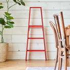 Een Hasegawa Lucano Ladder 3 Step Rood van Hasegawa staat tegen een witte houten planken muur naast een potplant. Houten stoelen en een eettafel zijn gedeeltelijk zichtbaar aan de rechterkant.
