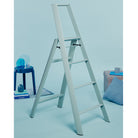 Een Hasegawa Lucano Ladder 4 Step in Dusty Mint van Hasegawa staat in een minimalistische kamer met blauwe muren, een bijpassende kruk met een lamp en boeken en een grijze poef met daarop een mok, boek en blauw kussen.