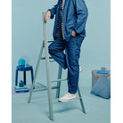 Een persoon in blauwe spijkerstof staat op een Hasegawa Lucano Ladder 4 Step Dusty Mint van Hasegawa in een pastelblauwe kamer met kussens, een bijzettafel en een mok. Het gezicht van de persoon is niet zichtbaar.