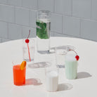 Vier kleurrijke drankjes in Hay Angle Longdrink Glazen van Hay staan op een witte tafel: één met munt, één met een schijfje sinaasappel, twee met kersen en één gewoon met schuim, allemaal tegen een betegelde witte achtergrond.