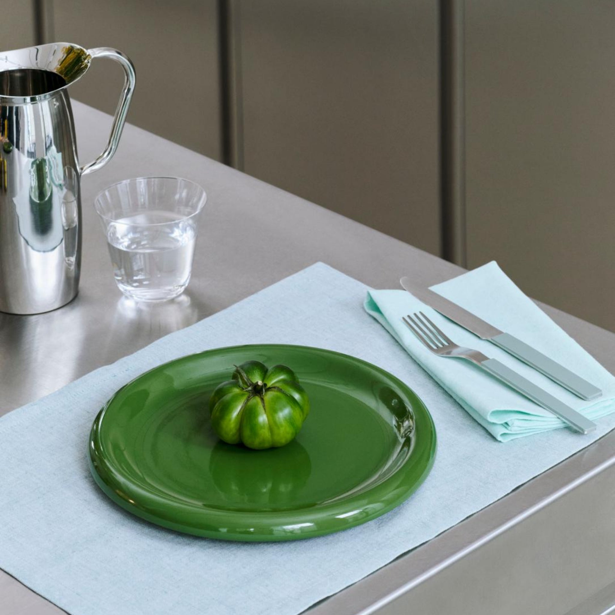 Een enkele groene tomaat rust op een Hay Barro Borden Groen D24 van Hay op een lichtblauwe placemat, met een vork en mes op een bijpassend servet vlakbij, plus een glas water en een zilveren kan op de roestvrijstalen tafel.