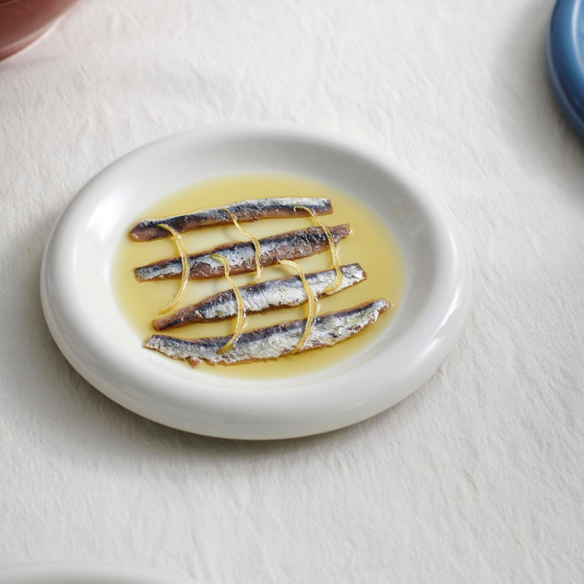 Vier ansjovisfilets zijn parallel gerangschikt op een Hay Barro Borden Off-White D18 bord van Hay, besprenkeld met olijfolie, bovenop een licht tafelkleed.