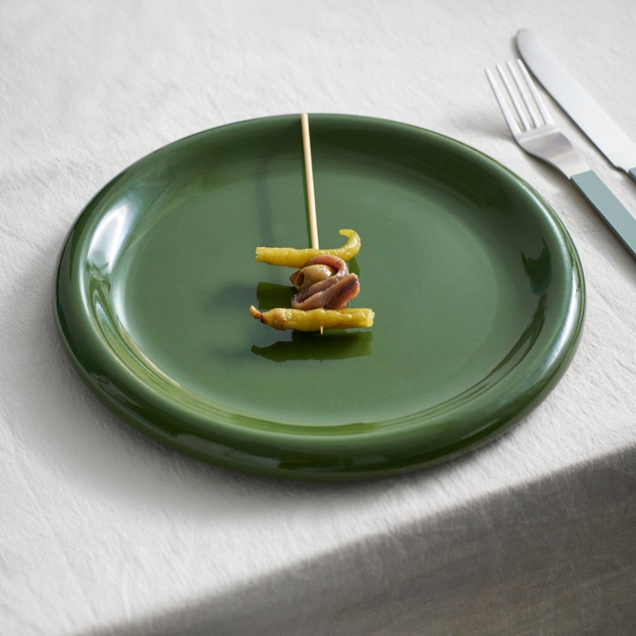 Een Hay Barro Borden Groen D24 bord van Hay heeft een spies van ingemaakte groenten en ansjovis in het midden, rustend op een wit tafelkleed naast een vork en mes.