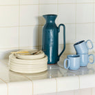 Een stapel crèmekleurige borden met gouden bestek staat op een betegeld aanrecht naast een Hay Barro Kan Donkerblauw 2L van Hay en drie lichtblauwe mokken, twee gestapeld. Een wit betegelde muur dient als achtergrond.