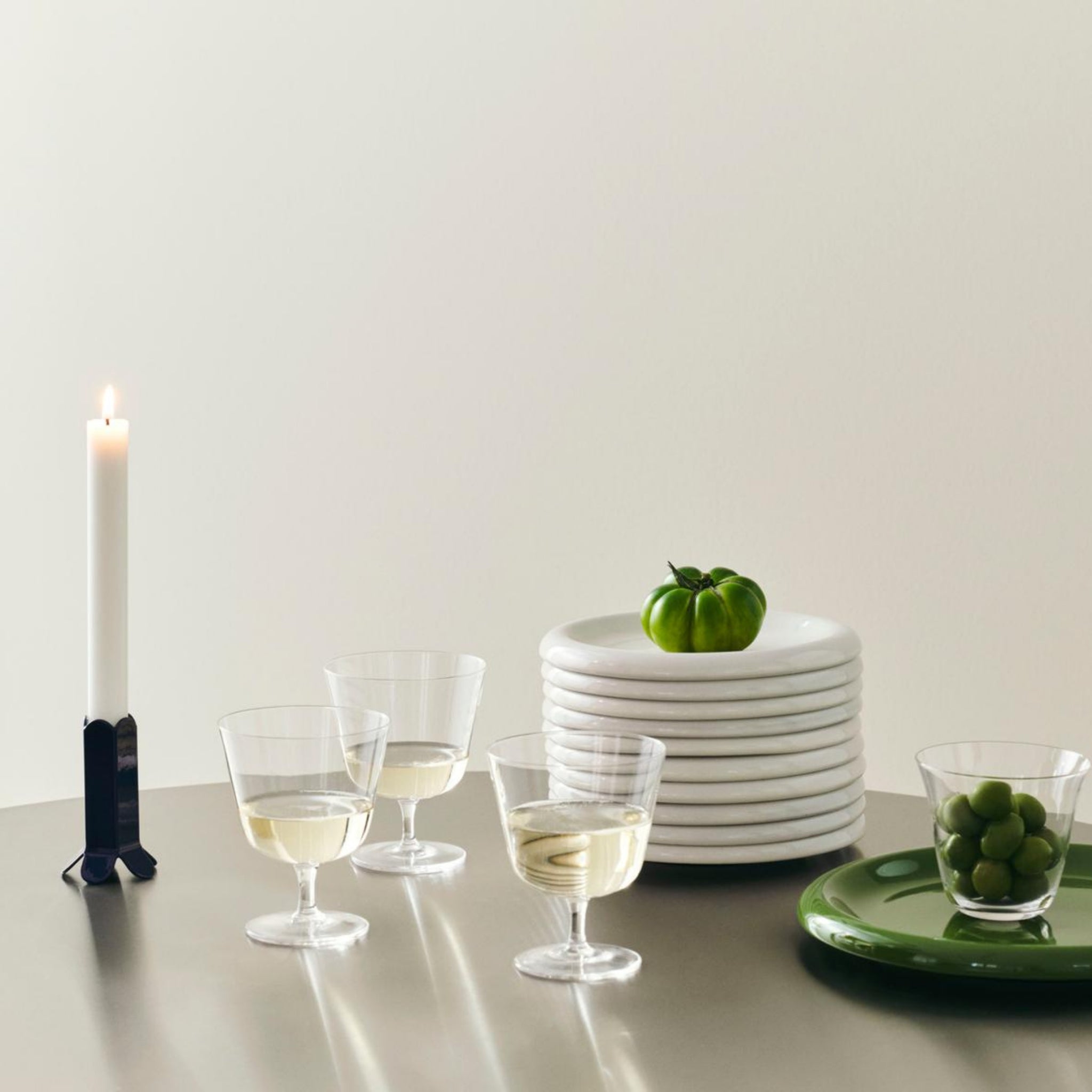 Een minimale tafelschikking bestaat uit drie glazen witte wijn, gestapelde witte borden met een groene tomaat, Hay Barro Borden Groen D24 van Hay met een schaal groene olijven en een brandende witte kaars in een zwarte houder.