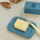 Een rechthoekig blok boter rust op de Hay Barro Boterschaal Donkerblauw van Hay, met een mes ernaast op een licht aanrecht. Het bijpassende blauwe deksel en sneetjes brood zijn zichtbaar op de achtergrond.