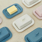 Rechthoekige boterschaaltjes van keramiek, waaronder de Hay Barro Boterschaal Roze van Hay, staan op een gespikkeld beige oppervlak in zachte pasteltinten. In één schaal past boter, terwijl de andere met deksel de terracotta servieslook aanvullen.