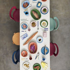 Een bovenaanzicht van een eettafel gedekt met Hay Barro Borden Groen D18 borden van Hay, geaccentueerd met terracotta borden, kleurrijk eten zoals vis, garnalen en groenten, en vier heldere stoelen op een grijze vloer.