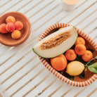 Een gestreepte schaal van keramiek uit de Barro-collectie staat op een tafel met witte latten, gevuld met meloen, citroenen, sinaasappels en mandarijnen. Ernaast staat de Hay Barro Ovale Schaal Terracotta 27/36 van Hay met abrikozen.