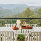 Op een witte buitentafel met bijpassende stoelen staat de Hay Barro Ovale Schaal Terracotta 27/36 van Hay, gevuld met gemengd fruit, een bord perziken en een kleine witte lamp, met uitzicht op groene bomen en bergen in de verte onder een bewolkte hemel.