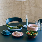 Een ronde tafel gedekt met pastelkleurige keramieken schaaltjes, een botervloot, Portugees servies, groenten in de Hay Barro Kom Groen D21 van Hay, glazen kopjes en een hoge kaars staat tegen een lichte stenen muur en donkere stoel.