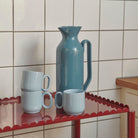 Een Hay Barro Kan Donkerblauw 2L keramische kan en drie gestapelde lichtblauwe mokken rusten op een metalen tafel met rode geschulpte randen, tegen een wit betegelde muur.