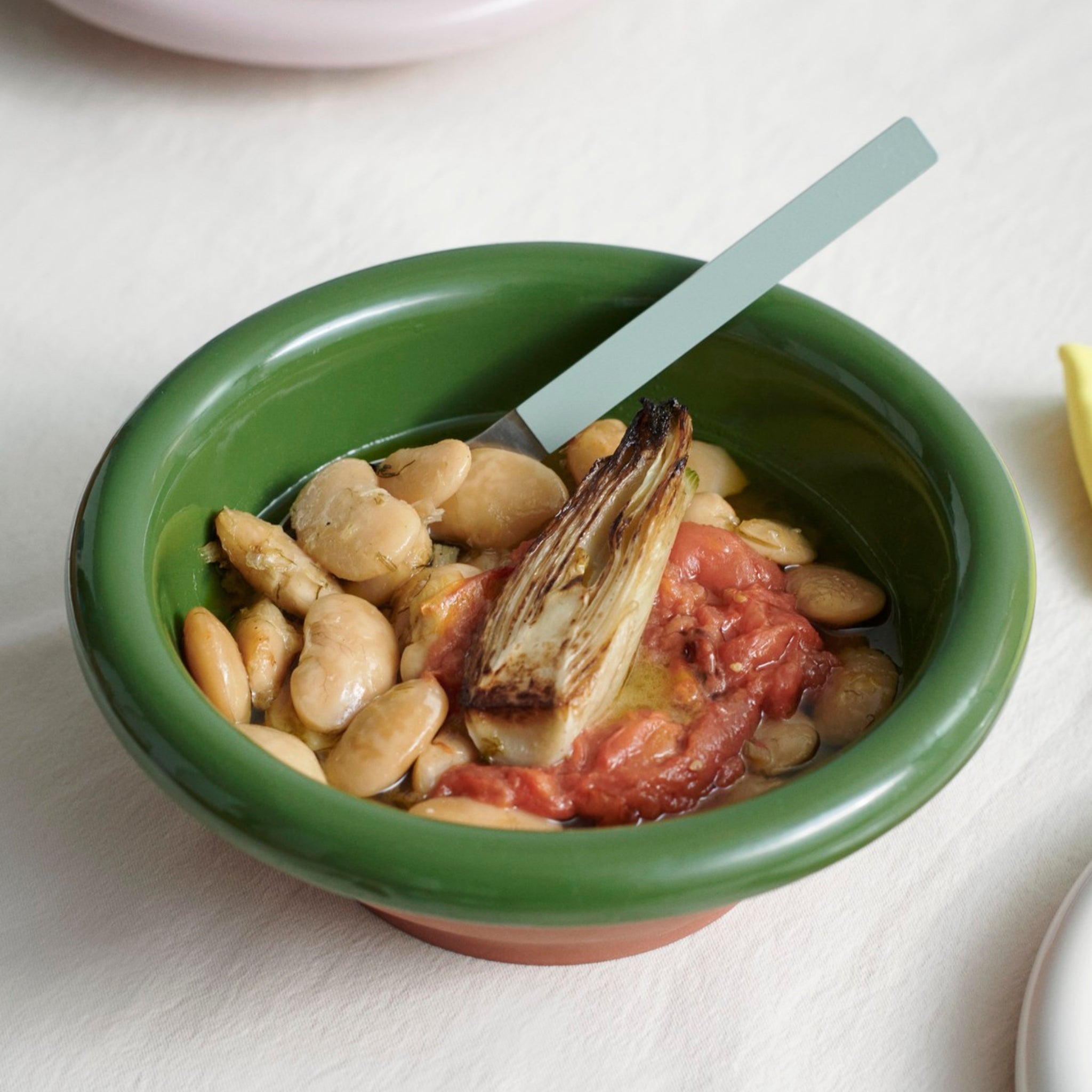 Een Hay Barro Kom Groen D21 van Hay, gevuld met witte bonen, een portie gestoofde tomaat en gegrilde andijvie, met een lepel in de kom.