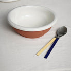 De Hay Barro Kom Off-White D26 kom van Hay staat op een wit kleed naast een lepel en vork met gele en blauwe handvatten.