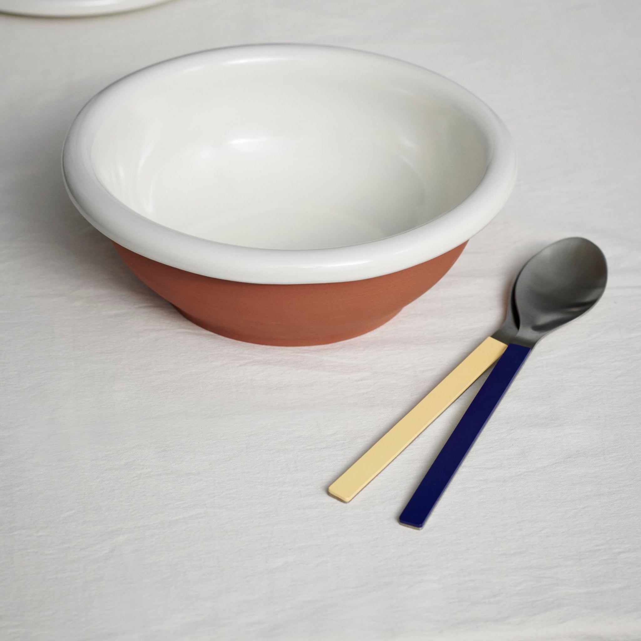 De Hay Barro Kom Off-White D26 kom van Hay staat op een wit kleed naast een lepel en vork met gele en blauwe handvatten.