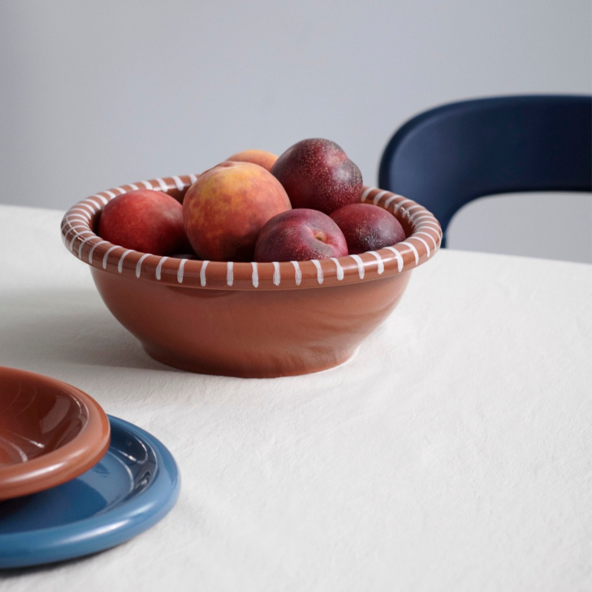 Een Hay Barro Kom Terracotta D26 kom met perziken en pruimen op een wit tafelkleed, met blauwe en bruine borden ernaast gestapeld en een donkerblauwe stoel gedeeltelijk zichtbaar op de achtergrond.